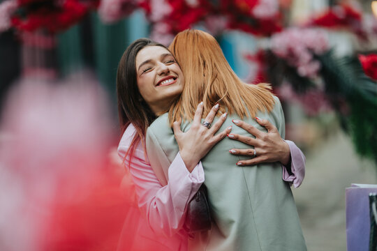 Two girls share a warm embrace while smiling on a flower-lined city street. The joyful friends hug each other amid colorful blossoms and urban surroundings.