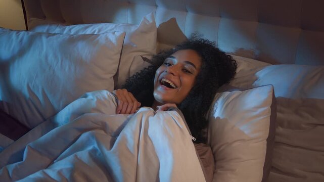 African American girl in bed pretending scary afraid covering duvet smiling laughing happy looking at camera girl female pretend frightened with nightmare darkness smile laugh at dark bedroom joke fun