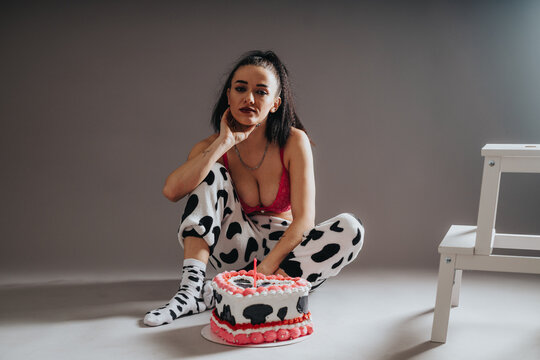 A young woman in cow-print pajama pants sits on the floor beside a decorated birthday cake. She poses casually in a pink bra and patterned socks for a relaxed birthday portrait.