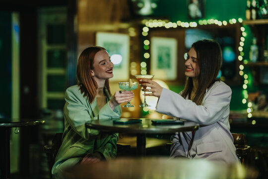Two young women sit at a bar smiling and clinking cocktail glasses. A casual nightlife scene of friends enjoying drinks and conversation.