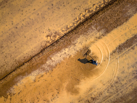 Aerial view of a farmer working in a muddy rice paddy field with visible ripples in the water and soil patterns in Bawen, Central Java, Indonesia.