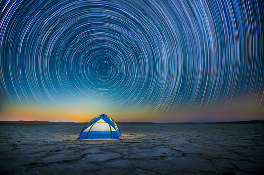 View of a blue and white tent set on a cracked desert floor under a vibrant night sky filled with circular star trails in the United Arab Emirates.