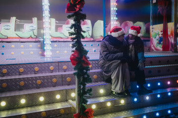 Fototapeta premium A couple wearing Santa hats sits close together on lit metal steps at a fairground. The romantic scene shows them sharing a quiet moment by colorful holiday lights.