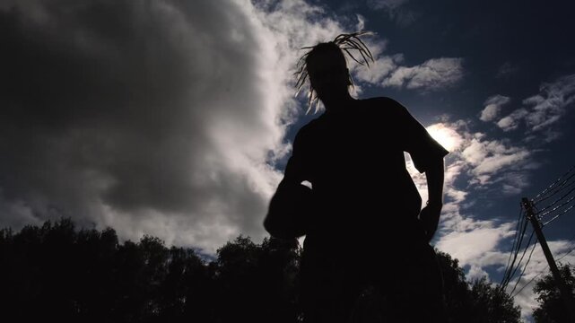 A silhouette of a man with long dreadlocks playing basketball, passing balls from hand to hand against the backdrop of the sky and bright sun