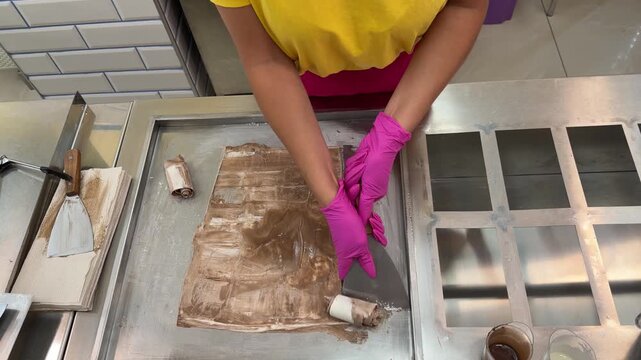 Gloved hands spread chocolate ice cream mixture flat on metal cold plate using spatula, small roll already formed beside it.