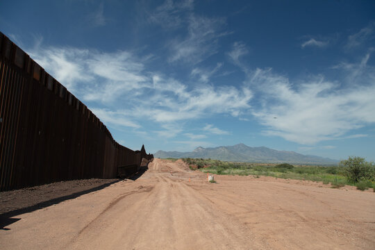 View of the metal border wall stretching along a dirt road in the desert landscape with mountains in the background in Naco, Arizona, United States.