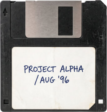 An old black floppy disk with a metallic shutter and a white paper label handwritten with blue ink text displaying project alpha and a date stamp from august nineteen ninety six