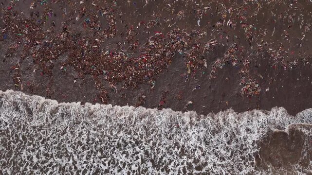 Aerial view of plastic waste covering the sandy beach with ocean waves washing debris ashore in Indonesia.