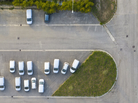 Aerial view of a parking lot with several white vans and cars parked on asphalt next to a green grass area in Fermo, Marche, Italy.