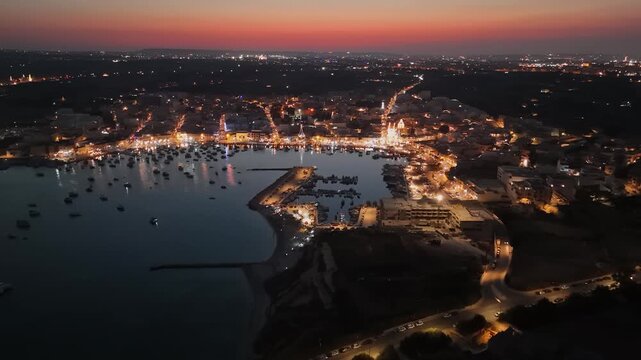 Marsaxlokk, Malta harbor with illuminated luzzu fishing boats and festival lights at dusk, reflecting on the waterfront during village festa celebrations in an aerial panorama.