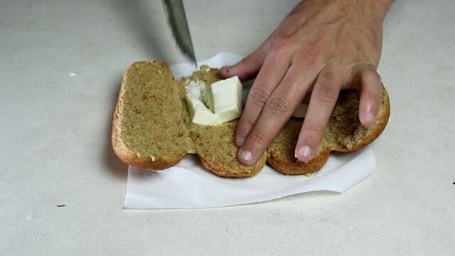 A close-up top-down view of male hands spreading soft butter onto a sliced brown bread roll using a knife. The food preparation takes place on a white napkin over a bright indoor kitchen counter.