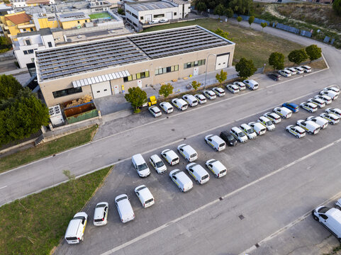 Aerial view of a commercial building with solar panels on the roof and a large fleet of white delivery vans parked in the lot in Fermo, Marche, Italy.