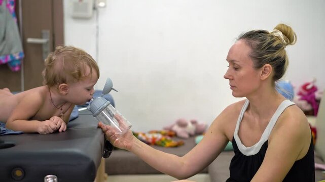 A woman is holding a baby and a bottle. The baby is laying on a table. The bottle is in the woman's hand