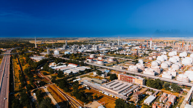Oil refinery tank farm beside highway under dark clouds. Drone view shows white tanks, process towers, pipelines, rail lines, warehouses and smoke near open fields.