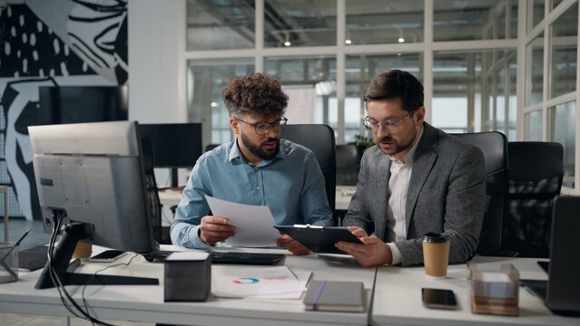 Two concentrated multiethnic business men working with documents discuss financial corporate budget planning income taxes diversity businessmen work talking looking in computer report talk in office