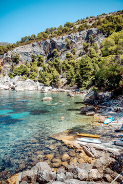 View of a rocky beach with crystal clear turquoise water, pine trees on the hillside, and paddleboards resting on the shore in Hvar, Split-Dalmatia County, Croatia.
