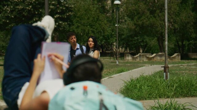 High school students engage in outdoor activities, with one drawing and others conversing. Young people enjoy their leisure time in a green park setting.