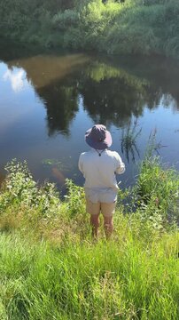 A fisherman with a fishing rod stands in the tall grass on a high riverbank on a sunny, clear summer day. Fisherman on the weekend. Hobbies, recreation and active recreation in summer