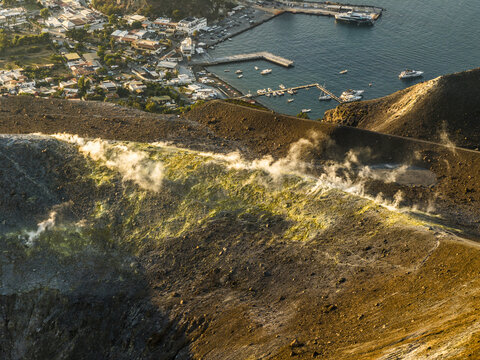 Aerial view of the steaming volcanic crater of Vulcano with sulfur deposits and rugged terrain overlooking the coastal town and harbor in Lipari, Sicily, Italy.