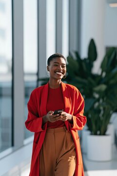 Smiling professional woman in red blazer walking with smartphone in modern office for business banner and background design