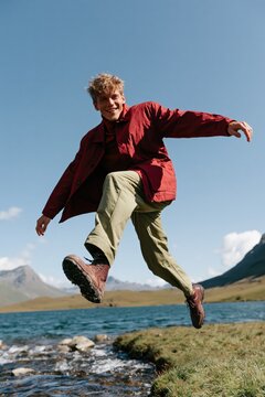 Young man jumping by mountain lake outdoors in a red jacket for adventure banner design and travel background