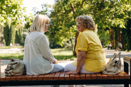 Two senior women engaged in conversation while sitting on a wooden bench in a lush green park, surrounded by trees and sunlight filtering through the leaves
