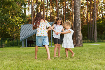 Fototapeta premium Three children dance and hold hands while standing on green grass in a park. The sun is low in the sky, creating a warm atmosphere. Trees surround them