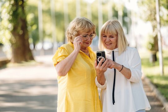 Two middle-aged women in casual attire, one in yellow and the other in white, smiling while looking at a smartphone in a sunny park setting