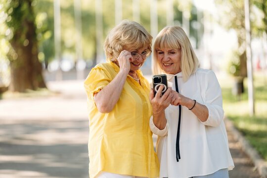 Two senior women with light hair and glasses sharing a moment while looking at a camera on a sunny day in a park with trees and pathways in the background