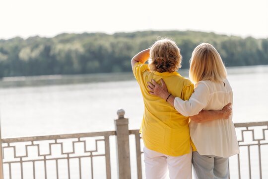 Two women standing together by a river, one wearing a yellow shirt and the other in a white blouse, enjoying the view with a railing in the foreground