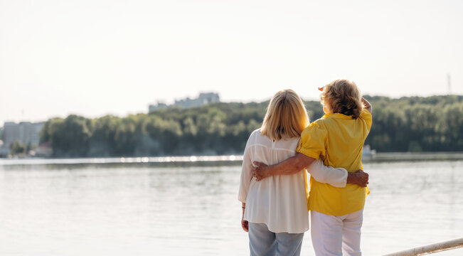 Two women standing by the water, one in a yellow shirt and the other in a white blouse, looking at the landscape with trees and buildings in the background