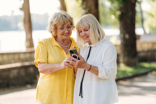 Two senior women with light hair wearing casual clothing smile while looking at a smartphone together in a sunny park setting near a body of water