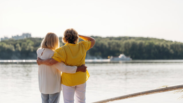 Two women standing by a river, one in a yellow shirt and the other in a white blouse, embracing while looking at the water and distant greenery