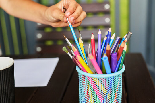 A person holds one pencil out of a cup filled with various colored pencils. A blank sheet of paper is on the table in a lively setting with green walls