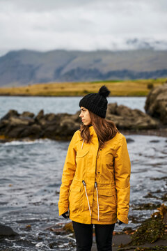White Woman in yellow jacket and black hat stands on rocky shoreline