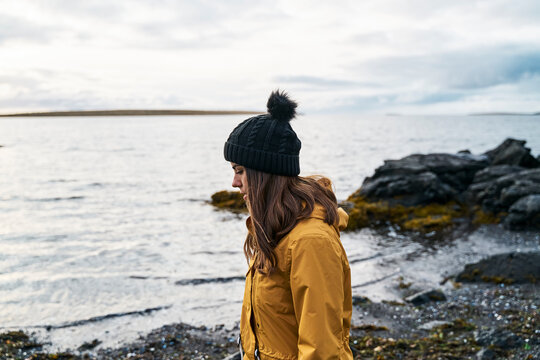 A white woman in a yellow jacket walks along a rocky shore