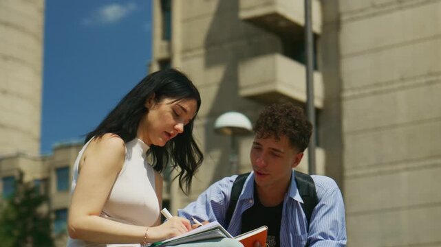 Two high school students are studying together outdoors, writing and discussing. They are collaborating on their schoolwork in a friendly setting.