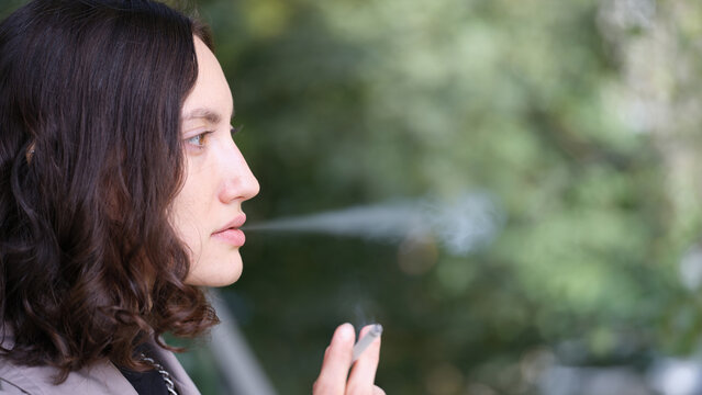 Side profile of young woman exhaling vapor from slim device outdoors, with soft natural light, blurred green background, calm expression, dark wavy hair, and close cropped composition emphasizing