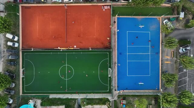 Aerial view of a sports complex featuring a red clay tennis court, a green soccer field, and a blue court with people playing in Porto San Giorgio, Marche, Italy.