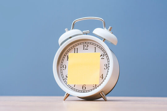 White alarm clock with a blank yellow sticky note on a wooden table, representing time management, reminders, deadlines, scheduling, productivity and daily planning.