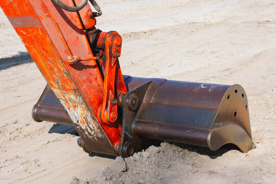 Close up of a heavy excavator bucket and hydraulic arm resting on sandy ground at a construction site, showing powerful machinery, earthmoving equipment, excavation and industrial work.