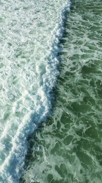 Aerial top view of turquoise ocean waves with white sea foam creating dynamic natural patterns and textures on moving seawater surface during a bright sunny summer day. Vertical video