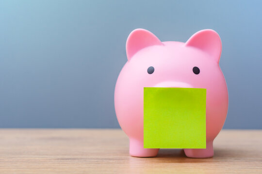 Pink piggy bank with a blank green note on a wooden table, representing savings, personal finance, budgeting, money management and financial planning with copy space.