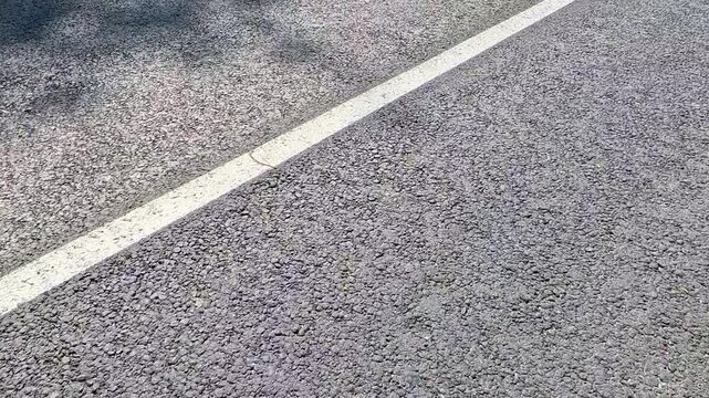 An empty gray asphalt road surface is shown close up from above, featuring a white dashed lane marking, with the camera moving steadily forward along the rough pavement texture in daylight.