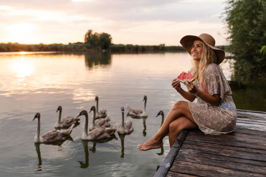 Woman Enjoys Summer by the River While Sitting on a Dock and Eating Watermelon Near Swans