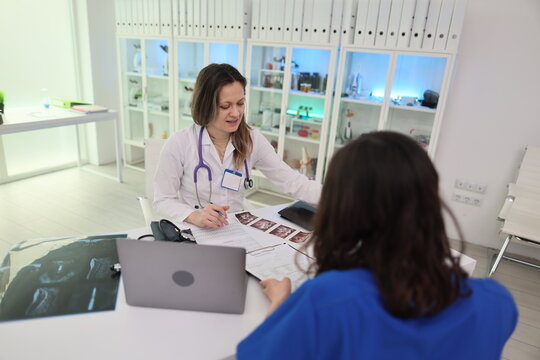 Smiling doctor in white coat and stethoscope reviews forms with nurse in blue scrubs during consultation at modern clinic desk, with laptop, clipboard, medical image, bright cabinets, clean