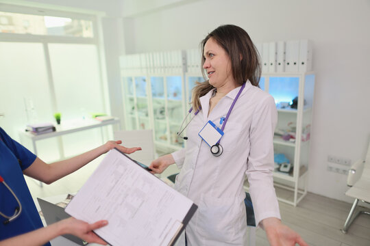 Female doctor in white coat smiles while greeting colleague during workplace handover inside bright medical office, with stethoscope, clipboard, shelves of binders, and soft daylight creating calm