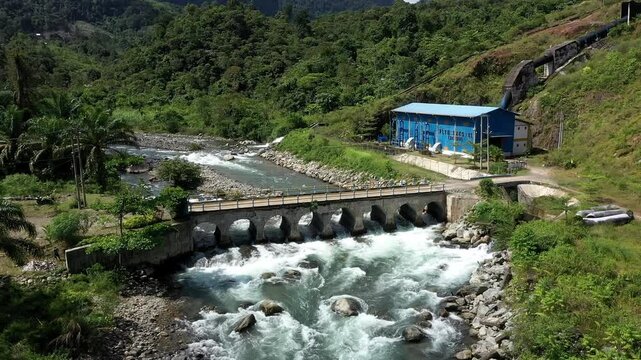 Aerial view of a small hydroelectric power plant situated on a rushing river with a stone bridge in the lush mountainous landscape of Kerinci Regency, Jambi, Indonesia.