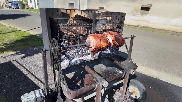 Whole pork shanks roasting slowly on a rotating spit outdoors, over glowing coals. Large pieces of pork rotate slowly on a metal spit over glowing embers and ashes, giving off a delicious aroma. 