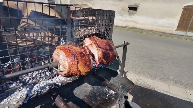 Whole pork shanks roasting slowly on a rotating spit outdoors, over glowing coals. Large pieces of pork rotate slowly on a metal spit over glowing embers and ashes, giving off a delicious aroma. 